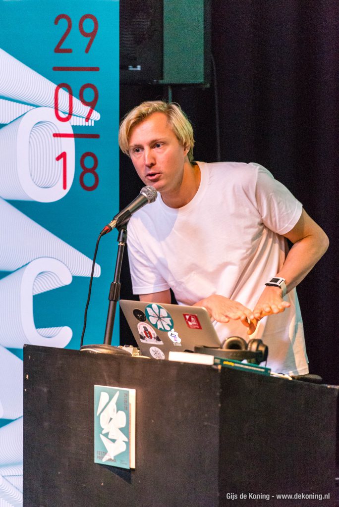 Lieven praat in een microfoon achter een spreekgestoelte. Achter hem een blauwe banner met de podcastfestival 2018 datum er op, voor hem zijn laptop en een koptelefoon