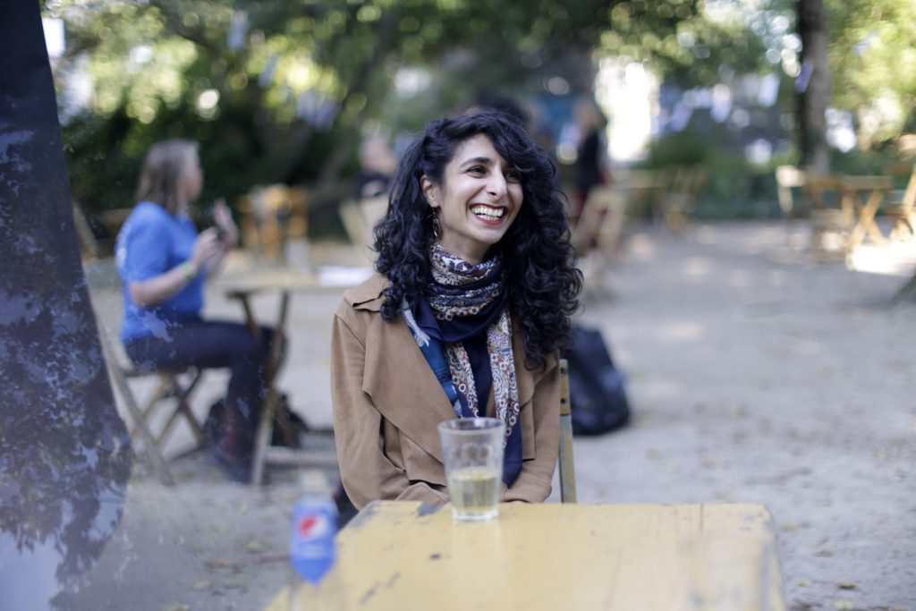 Mina zit in de tuin van de tolhuistuin aan een tafel en lacht heel leuk. Ze heeft lange donkere krullen en een sjahwltje om