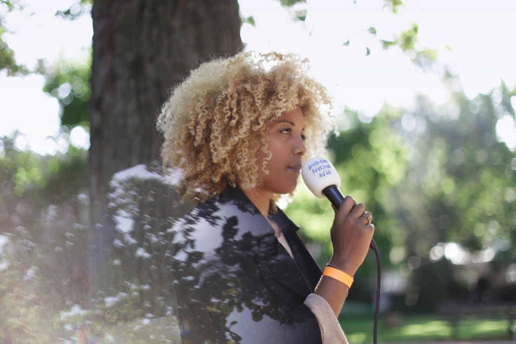 Foto van een vrouw met blonde afro en getinte huidskleur die in een microfoon met het logo van podcastfestival 2020 praat. Op de achtergrond zie je onscherp een boom en bladeren