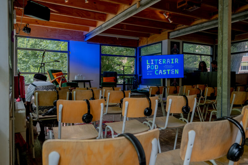 Foto van een zaal met daarin lege stoelen met ieder een koptelefoon er op. Op het podium een scherm met de vormgeving van het programma Literair Podcasten