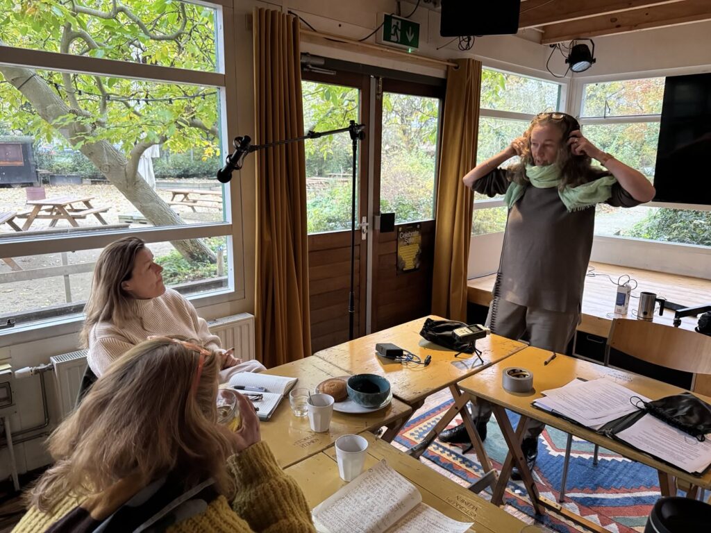 Foto van een podcastworkshop: twee mensen aan tafel met notitieboekjes, kopjes thee en opnameapparatuur, een derde persoon staat met een koptelefoon op. Op de achtergrond een binnenruimte met grote ramen en uitzicht op een terras.