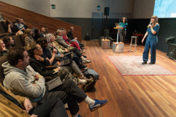Foto van een spreker op een houten podium. In de zaal zitten toeschouwers op stoelen.