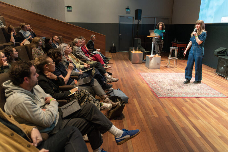 Foto van een spreker op een houten podium. In de zaal zitten toeschouwers op stoelen.