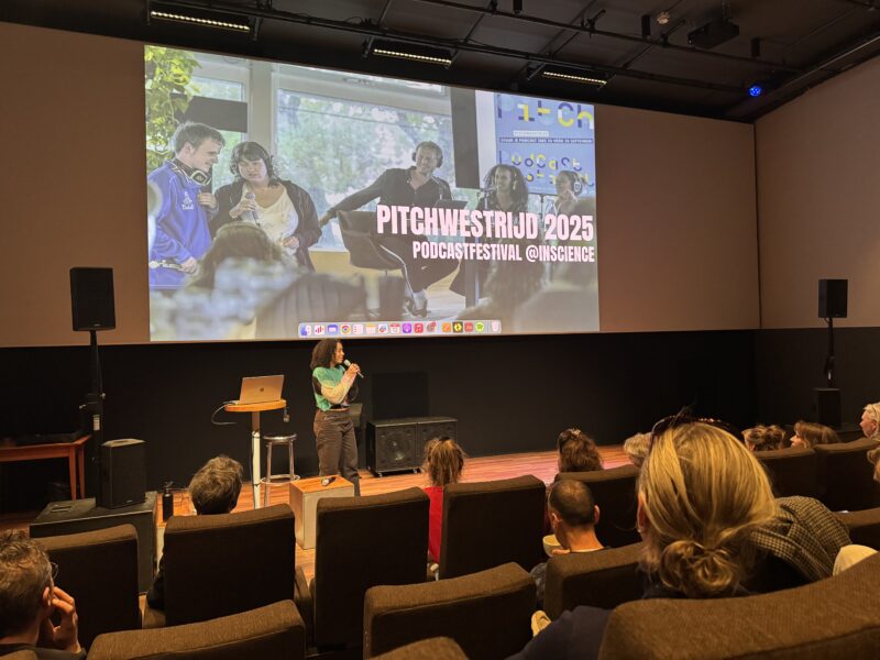 Foto van een presentator op een podium in een donkere zaal, voor een groot scherm waarop staat: “Pitchwedstrijd 2025 – Podcastfestival @InScience”. Op het scherm is een afbeelding te zien van mensen met microfoons in gesprek. In de zaal zitten toeschouwers op stoelen.