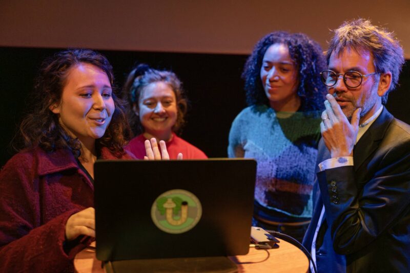Vier mensen staan rondom een laptop op een ronde tafel. Een jonge vrouw op de voorgrond praat en gebaart naar het scherm, terwijl de anderen geïnteresseerd meekijken. De sfeer is warm en gefocust, met kleurrijke podiumverlichting.