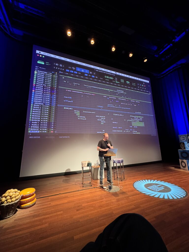 Zaal met houten vloer waarop een man achter een laptop staat met een microfoon in zijn handen die presenteert. Op het scherm achter hem is een projectie te zien van een audio-montageprogramma.