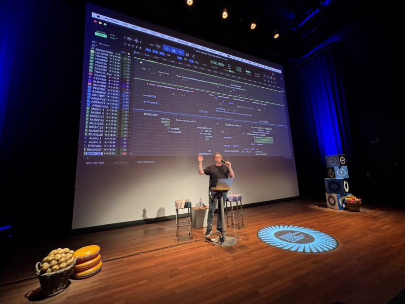 Zaal met houten vloer waarop een man achter een laptop staat met een microfoon in zijn handen die presenteert. Op het scherm achter hem is een projectie te zien van een audio-montageprogramma.