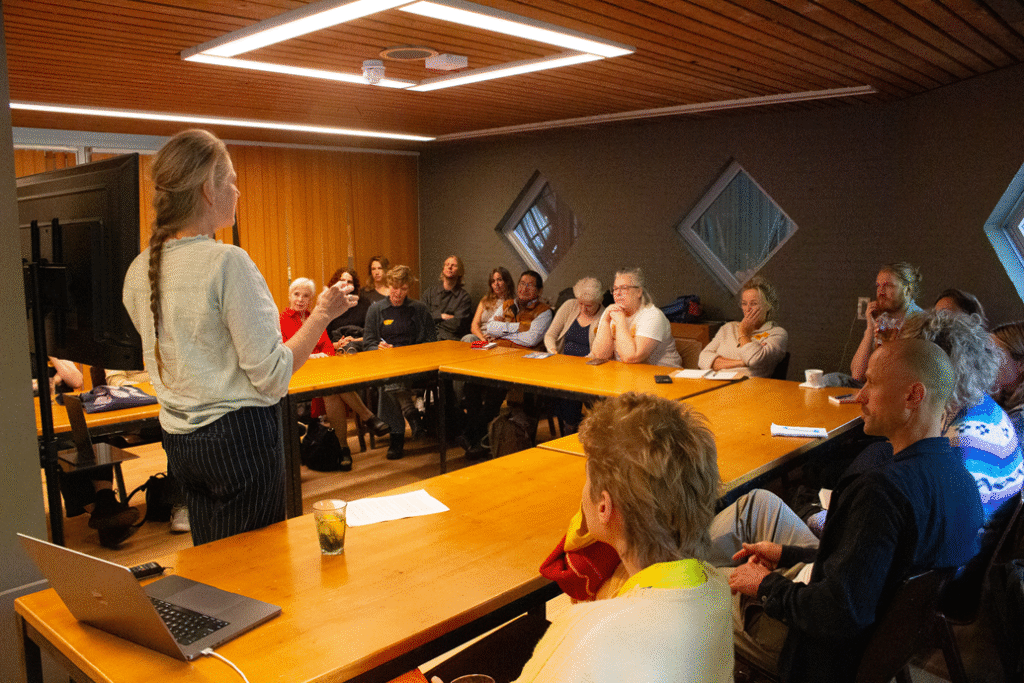 Foto van een kleine zaal met tafel in U-vorm waaraan mensen zitten. Ze luisteren naar een presentatie van iemand die in het midden voor een scherm staat te vertellen.
