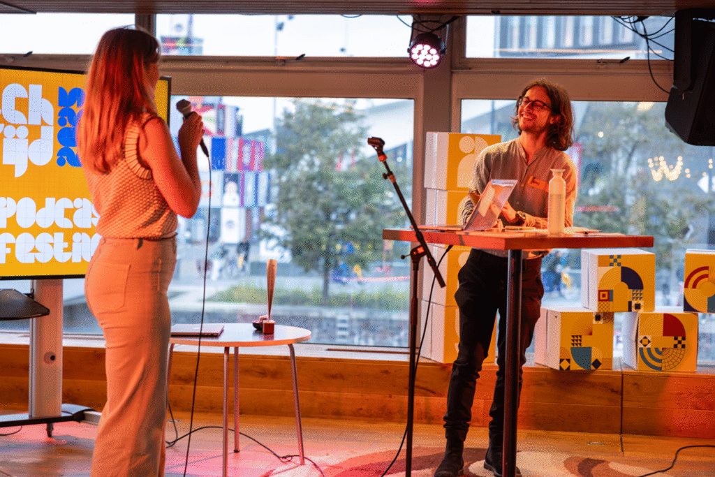 Foto van een podium waarop een spreker staat en Hans Jonker, de winnaar van de pitchwedstrijd.
