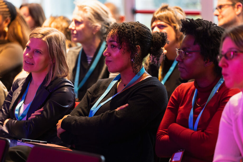 Een close-up van een aandachtig publiek tijdens het panel Podcastkritiek op OORZAKEN festival december 2025. De focus ligt op drie vrouwen op de voorgrond die geconcentreerd luisteren; ze zitten met hun armen over elkaar en kijken naar een spreker buiten beeld. Velen dragen lichtblauwe keycords. De belichting is warm met een zachte paarsroze gloed aan de rechterkant.