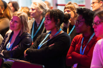 Een close-up van een aandachtig publiek tijdens het panel Podcastkritiek op OORZAKEN festival december 2025. De focus ligt op drie vrouwen op de voorgrond die geconcentreerd luisteren; ze zitten met hun armen over elkaar en kijken naar een spreker buiten beeld. Velen dragen lichtblauwe keycords. De belichting is warm met een zachte paarsroze gloed aan de rechterkant.