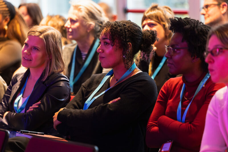 Een close-up van een aandachtig publiek tijdens het panel Podcastkritiek op OORZAKEN festival december 2025. De focus ligt op drie vrouwen op de voorgrond die geconcentreerd luisteren; ze zitten met hun armen over elkaar en kijken naar een spreker buiten beeld. Velen dragen lichtblauwe keycords. De belichting is warm met een zachte paarsroze gloed aan de rechterkant.