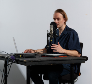 Tomas zit aan tafel achter microfoon en laptop