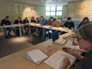 Foto van een groep mensen die in een zaal allemaal over een papier gebogen zitten en daar op werken. Er staan kopjes en laptops op tafel, op de muren hangen posters met anatomische tekeningen van de natuur.